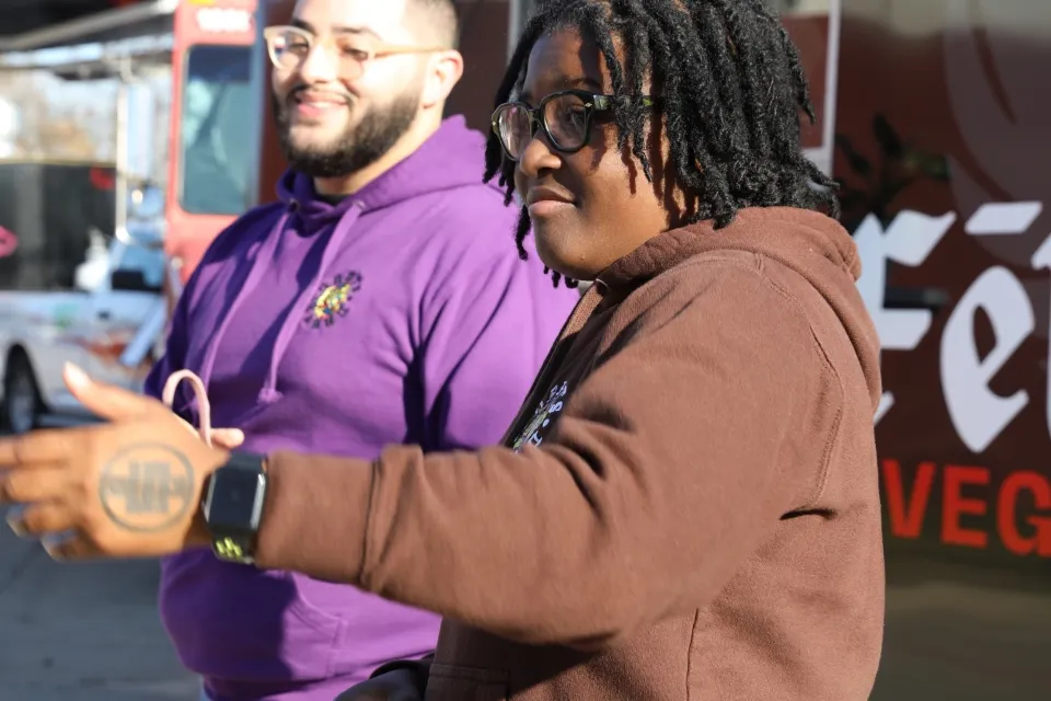 Black woman in brown sweatershirt and Black man in purple sweatshirt stand in front of food truck.